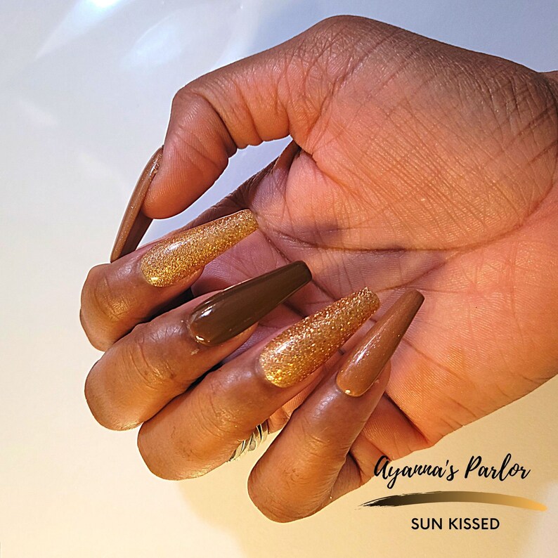 May include: Close-up of a hand with long, brown and gold glitter nails. The nails are in a coffin shape and are very long. The text "Ayanna's Parlor SUN KISSED" is visible in the bottom right corner of the image.