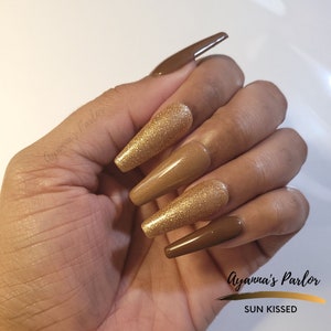 May include: Set of long, almond-shaped press-on nails with a brown and gold glitter ombre design. The nails are labeled "SUN KISSED" and "Ayanna's Parlor".