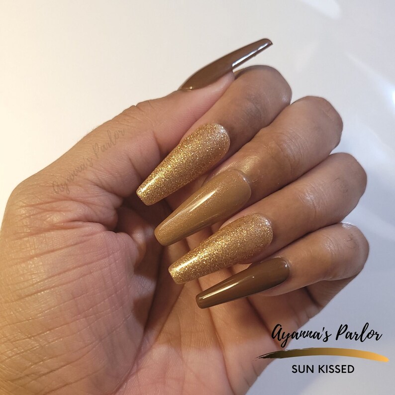 May include: Set of long, almond-shaped press-on nails with a brown and gold glitter ombre design. The nails are labeled "SUN KISSED" and "Ayanna's Parlor".