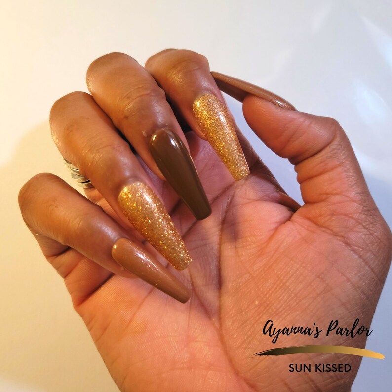 May include: Long, almond-shaped, brown and gold glitter press-on nails. The nails are shown on a hand with dark skin. The text "Ayanna's Parlor SUN KISSED" is visible in the bottom right corner of the image.