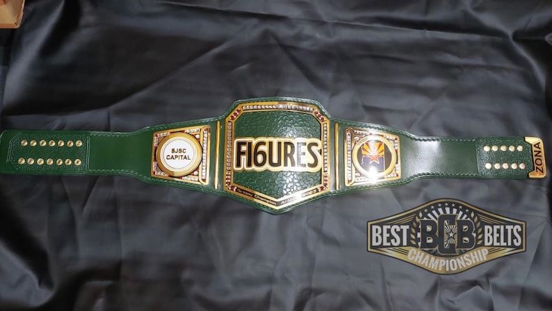 May include: A green leather championship belt with gold accents. The center plate features the word "FIGURES" in gold lettering. The side plates feature the words "S.J.S.C. CAPITAL" and a logo with a star and the text "ZONA". The belt is labeled "BEST B.C.B. BELTS CHAMPIONSHIP".