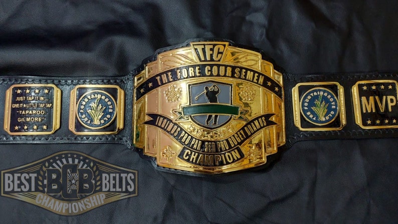May include: A gold and black championship belt with the text "TFC THE FORE COUR SEMEN" and "INDINOS PRO PAR, SER PRO BOGEY DRAMS CHAMPION". The belt has several side plates with text and logos.