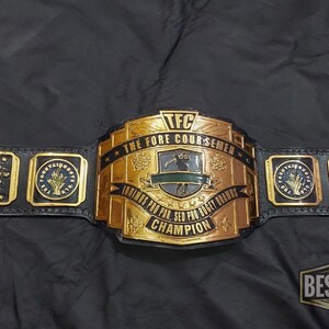 May include: A black leather championship belt with gold-colored metal plates. The belt features the text "TFC THE FORE COURSEMEN" and "FOREVER FOR PAR, SED FOR BIRDIE, CHAMPION". Additional plates display "MVP" and other text.