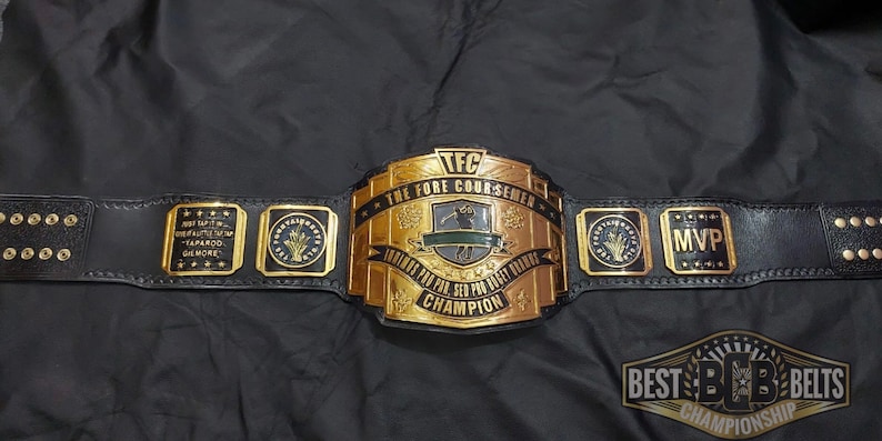 May include: A black leather championship belt with gold-colored metal plates. The belt features the text "TFC THE FORE COURSEMEN" and "FOREVER FOR PAR, SED FOR BIRDIE, CHAMPION". Additional plates display "MVP" and other text.