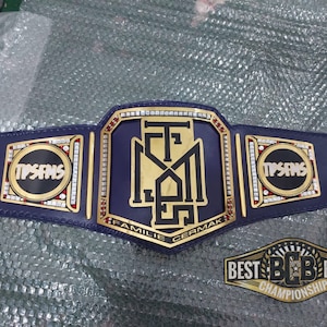 May include: A gold and blue championship belt with the text "TPSANS" on the side plates and "FAMILIE CERMAK" on the center plate. The belt is decorated with rhinestones and has a black leather strap.