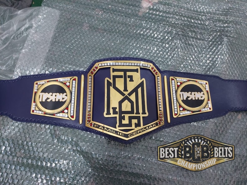 May include: A gold and blue championship belt with the text "TPSANS" on the side plates and "FAMILIE CERMAK" on the center plate. The belt is decorated with rhinestones and has a black leather strap.