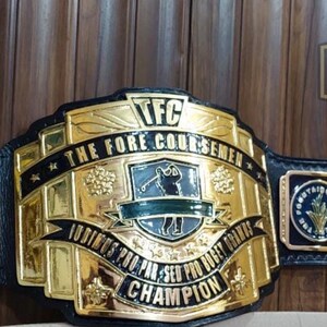 May include: A black leather championship belt with gold-colored metal plates. The central plate features the text "TFC THE FORE COURSEMEN CHAMPION". Additional plates include "MVP" and text that reads "JUST TAP IT IN GIVE IT A LITTLE TAP TAP TAPAROO GILMORE".