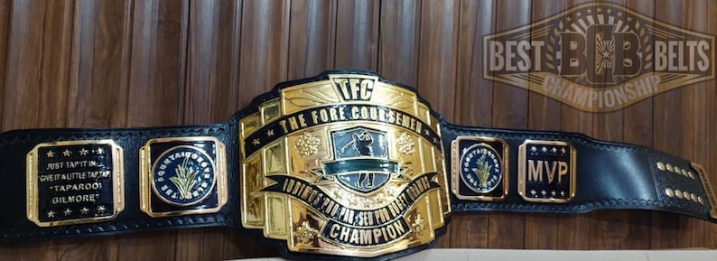 May include: A black leather championship belt with gold-colored metal plates. The central plate features the text "TFC THE FORE COURSEMEN CHAMPION". Additional plates include "MVP" and text that reads "JUST TAP IT IN GIVE IT A LITTLE TAP TAP TAPAROO GILMORE".