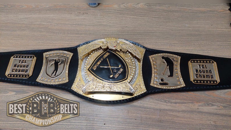 Puede incluir: Un cintur&oacute;n de campeonato dorado y negro con una correa de cuero negro y detalles en metal dorado. El cintur&oacute;n presenta un escudo central grande con un palo de golf y las palabras "Society of Leisure" y "Golf Club". El cintur&oacute;n tambi&eacute;n tiene dos escudos m&aacute;s peque&ntilde;os con una silueta de golfista y las palabras "TGL Round Champ".