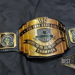 May include: A gold and black championship belt with the text "TFC THE FORE COURSEMEN CHAMPION" and "INDINOS PRO PAR, SED PRO BOGET URANTS CHAMPION". The belt has several side plates with text and designs. The belt is on a black surface.