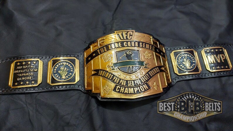 May include: A gold and black championship belt with the text "TFC THE FORE COURSEMEN CHAMPION" and "INDINOS PRO PAR, SED PRO BOGET URANTS CHAMPION". The belt has several side plates with text and designs. The belt is on a black surface.