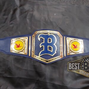 May include: A blue and gold championship belt with a large blue "B" in the center. The belt has two side plates with a yellow circle and a red bird. The text "BEST BCB BELTS CHAMPIONSHIP" is on the bottom of the belt.