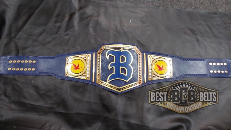 May include: A blue and gold championship belt with a large blue "B" in the center. The belt has two side plates with a yellow circle and a red bird. The text "BEST BCB BELTS CHAMPIONSHIP" is on the bottom of the belt.