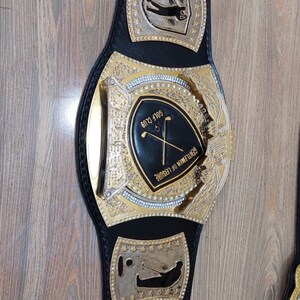 Puede incluir: Un cintur&oacute;n de campeonato de cuero negro con detalles dorados. La placa central presenta un escudo negro con el texto "GOLF CLUB" y "GENTLEMEN OF LEISURE". El cintur&oacute;n tiene dos placas laterales con el texto "TGL Round Champ" y una silueta de golfista.