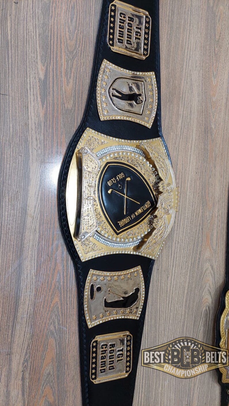 Puede incluir: Un cintur&oacute;n de campeonato de cuero negro con detalles dorados. La placa central presenta un escudo negro con el texto "GOLF CLUB" y "GENTLEMEN OF LEISURE". El cintur&oacute;n tiene dos placas laterales con el texto "TGL Round Champ" y una silueta de golfista.