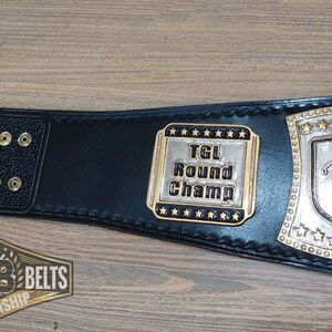 Puede incluir: Cintur&oacute;n de campeonato de cuero negro con detalles dorados. El cintur&oacute;n presenta una placa plateada con un golfista y el texto "TGL Round Champ".