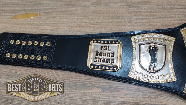 Puede incluir: Cintur&oacute;n de campeonato de cuero negro con detalles dorados. El cintur&oacute;n presenta una placa plateada con un golfista y el texto "TGL Round Champ".