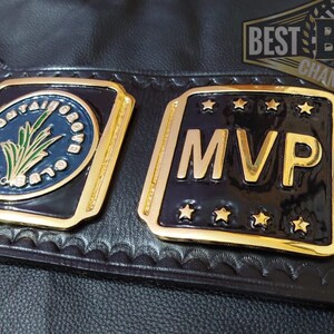 May include: Close-up of a black leather championship belt with gold and black plates. One plate reads "MVP" with gold stars. The other plate has "THE MOUNTAIN GROVE CLUB" in a blue circle with a green plant design. The belt has the text "BEST BOB BELTS CHAMPIONSHIP".