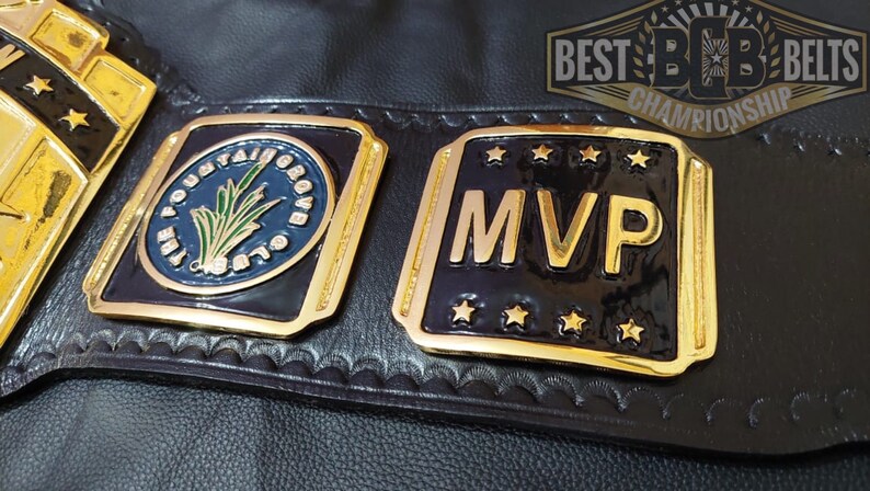 May include: Close-up of a black leather championship belt with gold and black plates. One plate reads "MVP" with gold stars. The other plate has "THE MOUNTAIN GROVE CLUB" in a blue circle with a green plant design. The belt has the text "BEST BOB BELTS CHAMPIONSHIP".