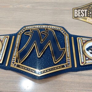 May include: A blue and gold championship belt with a large "M" in the center, surrounded by rhinestones. The belt also features two smaller side plates with a white panther logo. The text "Morton Panthers" is written on the belt.