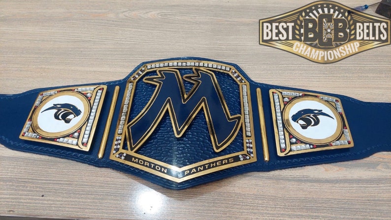 May include: A blue and gold championship belt with a large "M" in the center, surrounded by rhinestones. The belt also features two smaller side plates with a white panther logo. The text "Morton Panthers" is written on the belt.