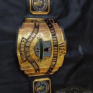 May include: A gold and black championship belt with the text "TFC" and "The Fore Coursemen". The belt has multiple gold plates with text and star details. The belt is on a black surface.