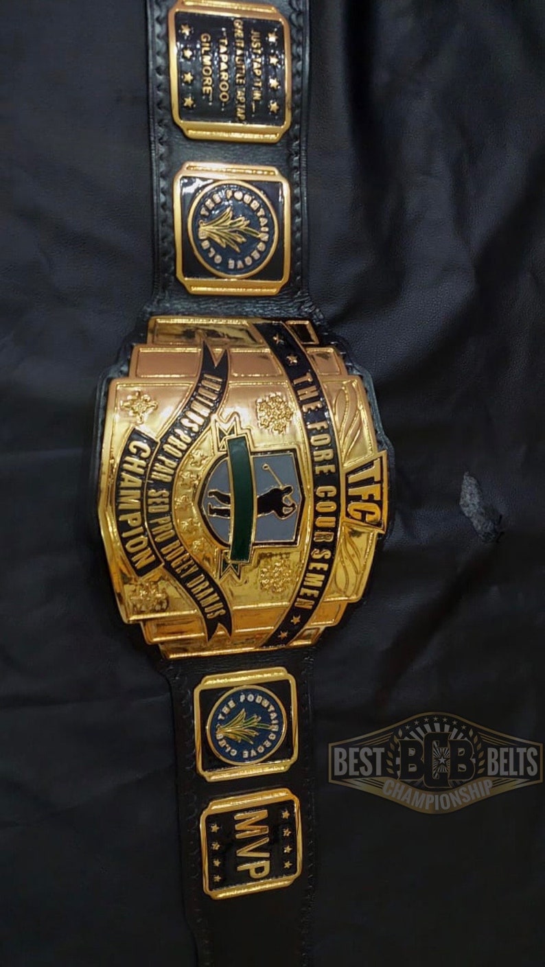 May include: A gold and black championship belt with the text "TFC" and "The Fore Coursemen". The belt has multiple gold plates with text and star details. The belt is on a black surface.