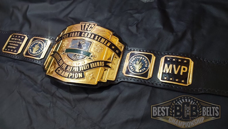 May include: A gold and black championship belt with the text "TFC THE FORE COUR SEMEN" and "CHAMPION". The belt has gold plates with the word "MVP" and other text. The belt is on a black surface.
