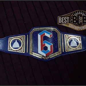 May include: A blue wrestling championship belt with gold accents and a large white, red, and blue number "6" in the center. The belt has two side plates with a white silhouette of a building on a blue background. The belt buckle reads "BEST BCB BELTS CHAMPIONSHIP".
