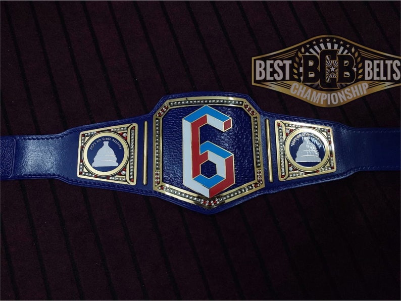May include: A blue wrestling championship belt with gold accents and a large white, red, and blue number "6" in the center. The belt has two side plates with a white silhouette of a building on a blue background. The belt buckle reads "BEST BCB BELTS CHAMPIONSHIP".