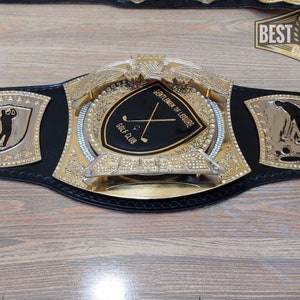 Puede incluir: Un cintur&oacute;n de campeonato de cuero negro con detalles dorados y pedrer&iacute;a. La placa central presenta un escudo negro con palos de golf cruzados y el texto "Gentlemen of Leisure" y "Golf Club". Las placas laterales presentan una silueta de golfista y el texto "TGL Round Champ".