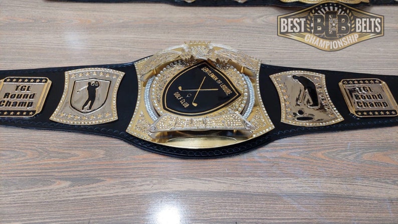 Puede incluir: Un cintur&oacute;n de campeonato de cuero negro con detalles dorados y pedrer&iacute;a. La placa central presenta un escudo negro con palos de golf cruzados y el texto "Gentlemen of Leisure" y "Golf Club". Las placas laterales presentan una silueta de golfista y el texto "TGL Round Champ".