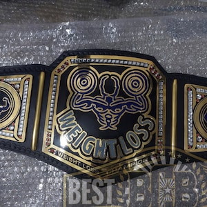 May include: A gold and black championship belt with rhinestones. The center plate features a weightlifting design with the text "WEIGHT LOSS CHAMPION".
