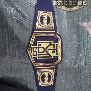 May include: A gold championship belt with a blue strap and gold plates. The center plate features a black and gold logo with the text "FAMILY CERMAK" and "SDX" inside a gold frame. The side plates feature a red and white circle with the text "TRISHES" in black.