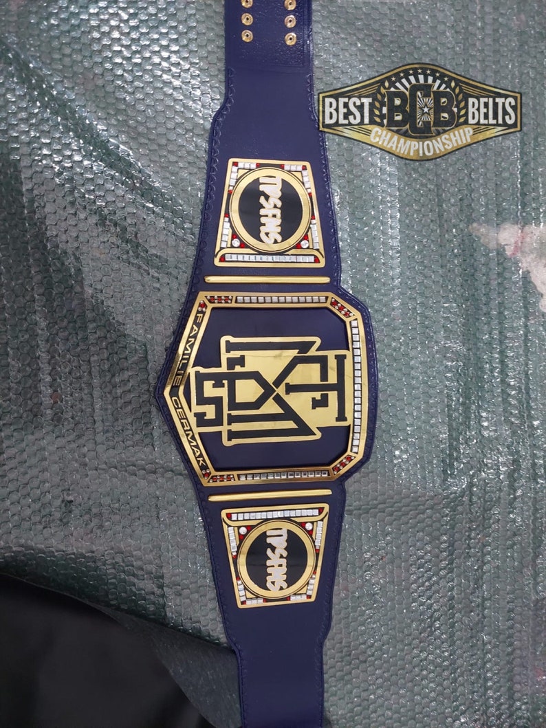 May include: A gold championship belt with a blue strap and gold plates. The center plate features a black and gold logo with the text "FAMILY CERMAK" and "SDX" inside a gold frame. The side plates feature a red and white circle with the text "TRISHES" in black.