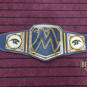 May include: A blue and gold championship belt with a large "M" in the center and a panther logo on each side. The belt is made of leather and has a gold buckle. The text "Morton Panthers" is printed on the belt.