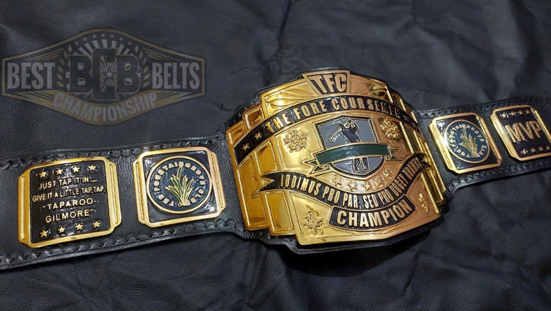 May include: A gold and black championship belt with the text "BEST BELTS CHAMPIONSHIP" at the top. The belt has multiple plates with text including "TFC THE FORE COURSE SERIES" and "MVP". The belt also has the text "JUST TAP IT IN GIVE IT A LITTLE TAP TAP TAPAROO GILMORE".