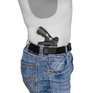 May include: A black handgun in a leather holster is attached to a black belt worn around a person's waist. The person is wearing blue jeans and a white tank top.