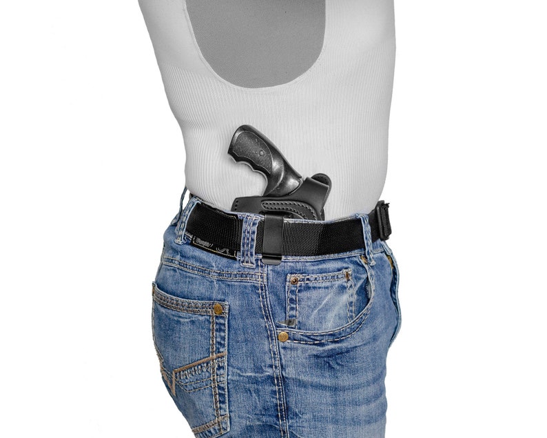 May include: A black handgun in a leather holster is attached to a black belt worn around a person's waist. The person is wearing blue jeans and a white tank top.