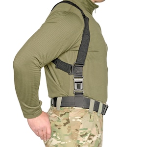 May include: A black tactical harness system featuring adjustable nylon webbing straps, heavy-duty plastic buckles, and reinforced stitching. The harness is worn over an olive green long-sleeve textured shirt and is attached to a black utility belt.