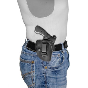May include: A black leather holster with a silver handgun inside. The holster is attached to a brown belt and worn on a person's waist.