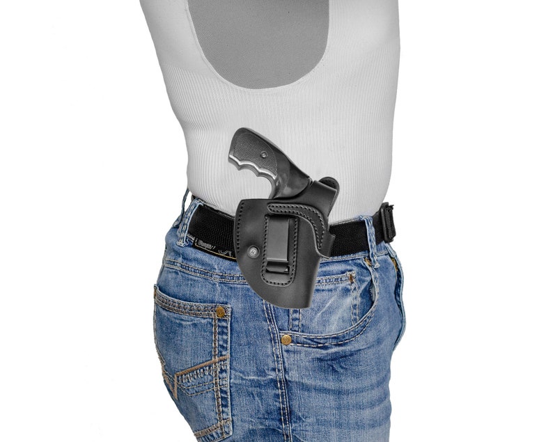 May include: A black leather holster with a silver handgun inside. The holster is attached to a brown belt and worn on a person's waist.