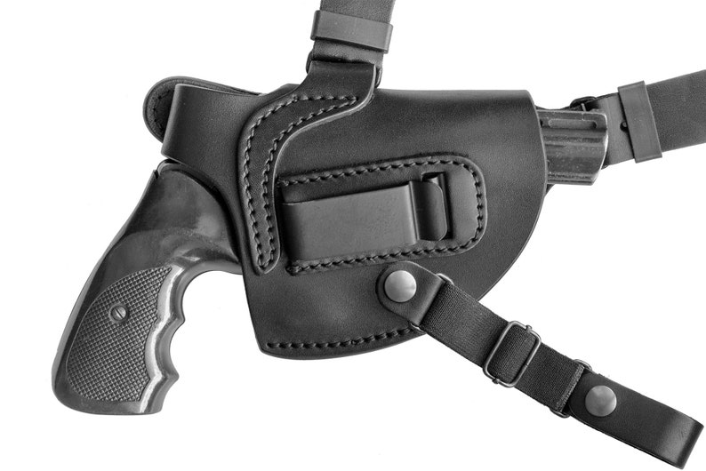 May include: A black leather shoulder holster with a black revolver inside. The holster has a strap that goes over the shoulder and a clip that attaches to the belt.
