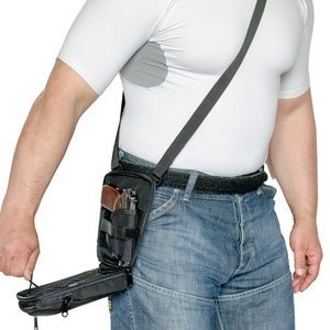 Puede incluir: Un holster táctico de hombro negro con una pistola en su interior. El holster tiene un bolsillo con cremallera en la parte inferior. El holster lo lleva puesto una persona que lleva una camisa blanca y unos vaqueros azules.