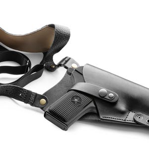 Puede incluir: Funda de hombro de cuero negro con una pistola negra a juego. La funda tiene una correa que va sobre el hombro y alrededor de la espalda. La pistola es parcialmente visible, con un emblema de estrella.