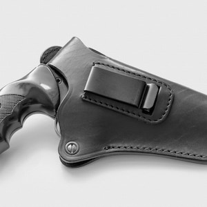 Può includere: Un fodero in pelle nera con una clip in metallo per una pistola. Il fodero è progettato per essere indossato su una cintura.