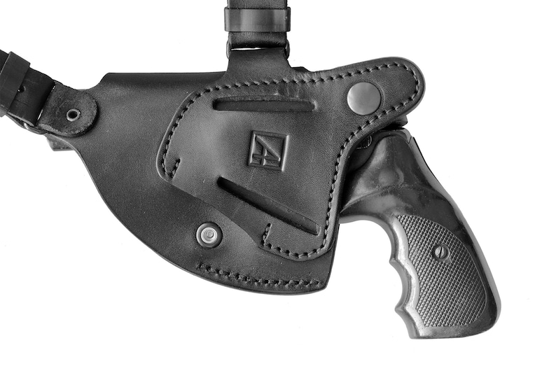 May include: A black leather holster with a shoulder strap for a small revolver. The holster has a snap closure and a metal button on the flap. The revolver has a black handle with a textured grip.