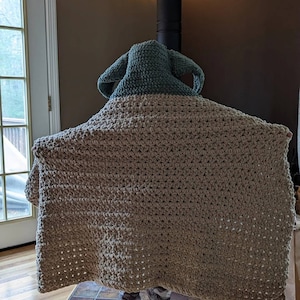 Puede incluir: Una manta de ganchillo hecha a mano en color beige y verde salvia, con una capucha con orejas puntiagudas. La manta est&aacute; drapeada sobre una persona, ocultando su cuerpo. La manta parece estar hecha de un hilo grueso, con una puntada texturizada.