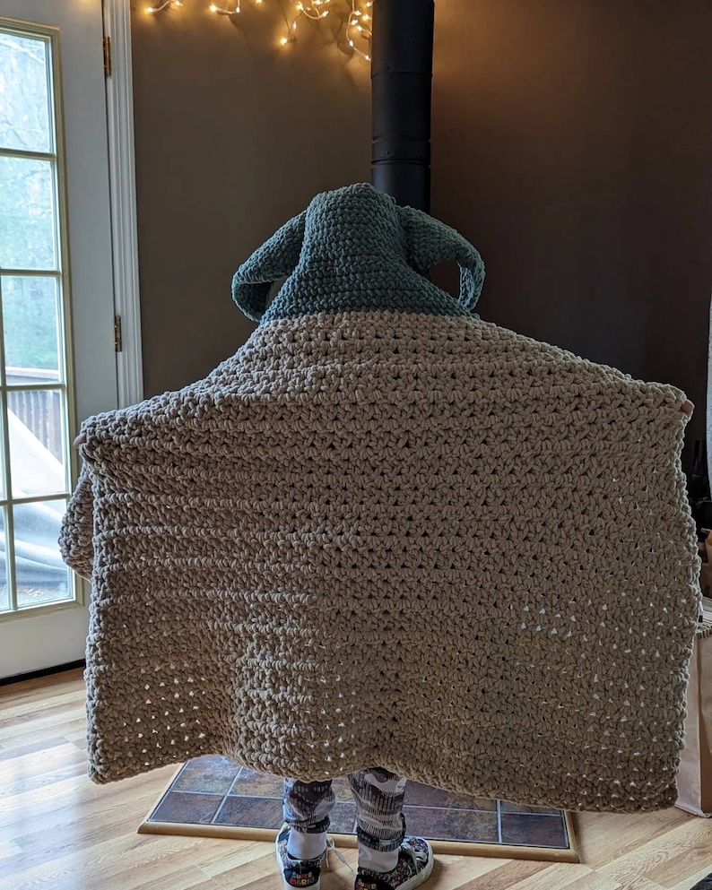 Puede incluir: Una manta de ganchillo hecha a mano en color beige y verde salvia, con una capucha con orejas puntiagudas. La manta est&aacute; drapeada sobre una persona, ocultando su cuerpo. La manta parece estar hecha de un hilo grueso, con una puntada texturizada.