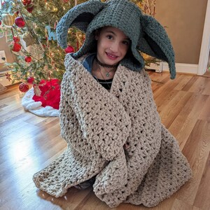 Puede incluir: Un ni&ntilde;o lleva un gorro verde tejido con grandes orejas puntiagudas y est&aacute; envuelto en una manta beige tejida. El gorro se asemeja a un personaje de una popular franquicia de ciencia ficci&oacute;n. La manta es gruesa y texturizada. Un &aacute;rbol de Navidad decorado est&aacute; en el fondo.